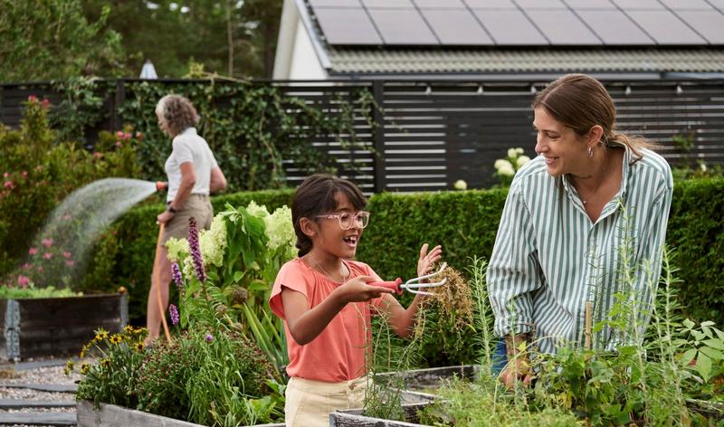 Mamma och barn tar hand om planteringar i trädgården. Solpaneler på huset i bakgrunden.